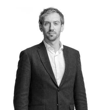 Gregory stands and looks at the camera. He wears a black suit and white shirt.