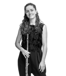 Marta stands and smiles with her flute held down. She wears a smart black jumpsuit.