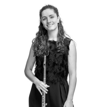 Marta stands and smiles with her flute held down. She wears a smart black jumpsuit.