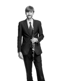 William stands with his clarinet. He smiles and wears a dark suit and tie.