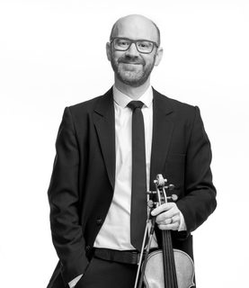Gordon stand with his violin. He smiles and wears a black suit and tie. He also wears glasses.