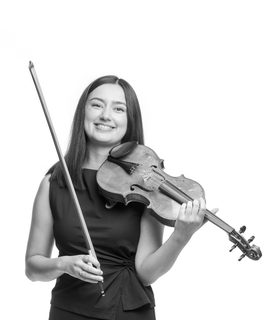 Michelle smiles to the camera as her violin rests under her neck and bow upright in the other hand. She wears a dark top and trousers.