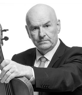 Steve looks to the camera and holds out the viola in front of him. He wears a dark jacket and white shirt with tie.