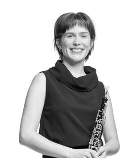 Katherine stands and holds her Oboe - she smiles and wears a dark top.