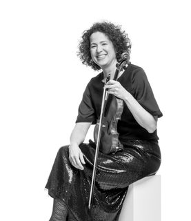 Rachel smiles and sits to the side holding her violin and bow. She wears a dark shimmering dress.