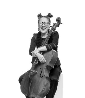 Su-a laughs. She sits with her cello resting back onto her. She wears her hair in space buns, wears glasses and a dark outfit.