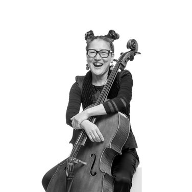 Su-a laughs. She sits with her cello resting back onto her. She wears her hair in space buns, wears glasses and a dark outfit.