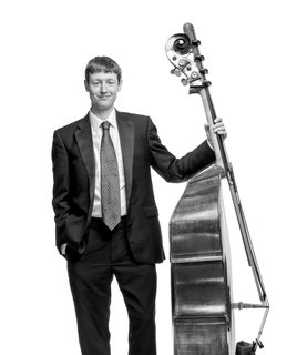 Jamie stands with his double bass up but facing to the side. He smiles and wears a dark suit and tie.