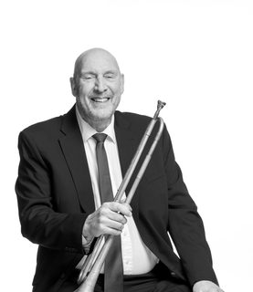 Shaun sits and laughs holding his trumpet. He wears a dark suit.