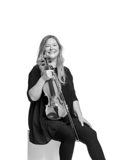 Amira sits holding her violin upright. She wears smart black top, trousers and shoes.