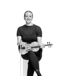 Hatty sits with her legs crossed, holding her violin across her lap. She smiles and wears a dark top, dark trousers and dark shoes..