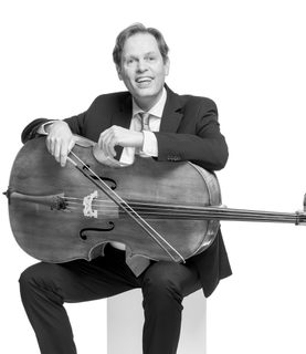Eric smiles and sits with the cello lying across his lap. He wears a dark suit and white shirt with tie.