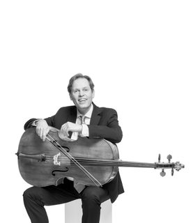 Eric smiles and sits with the cello lying across his lap. He wears a dark suit and white shirt with tie.