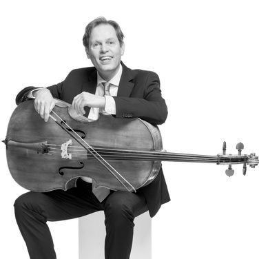 Eric smiles and sits with the cello lying across his lap. He wears a dark suit and white shirt with tie.