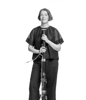 Cerys stands, resting her bassoon upright. She wears a black top and trousers.