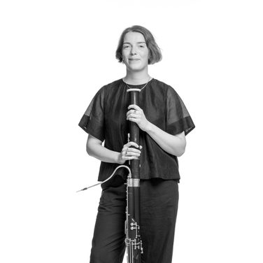 Cerys stands, resting her bassoon upright. She wears a black top and trousers.
