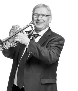 Peter laughs as he holds his trumpet out in front of him ready to play. He wears glasses and a black suit.
