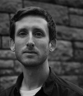 Daniel looks to the camera. The photo is black and white. He has short heair and wears a tshirt and shirt.