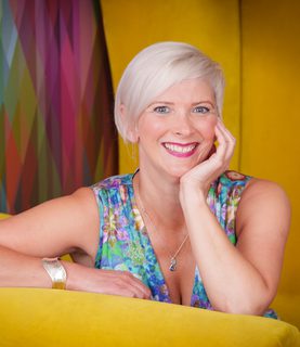 Emma smiles. She sits on a yellow chairs, resting her chin on her arm. She wears a blue, colourfullly patterened dress and has a short blonde bob.