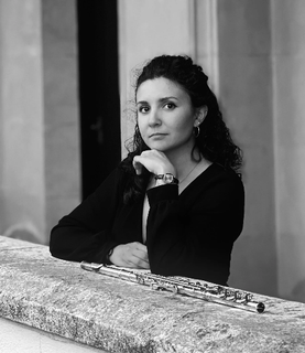 Alba Vinti rests her elbow on a ledge where her flute lies. She looks to the camera. Black and white photograph.