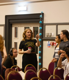 Sally talks to young people and points in the distance. She wears an SCO branded black top.
