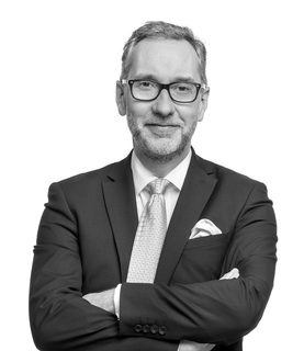 Gavin stands with his arms crossed. He wears glasses and a black suit and light tie.