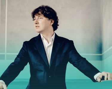 Maxim wears a suit and looks to the left of screen