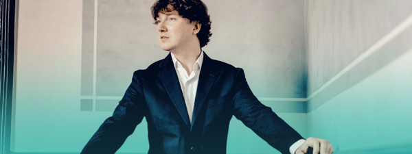 Maxim wears a suit and looks to the left of screen