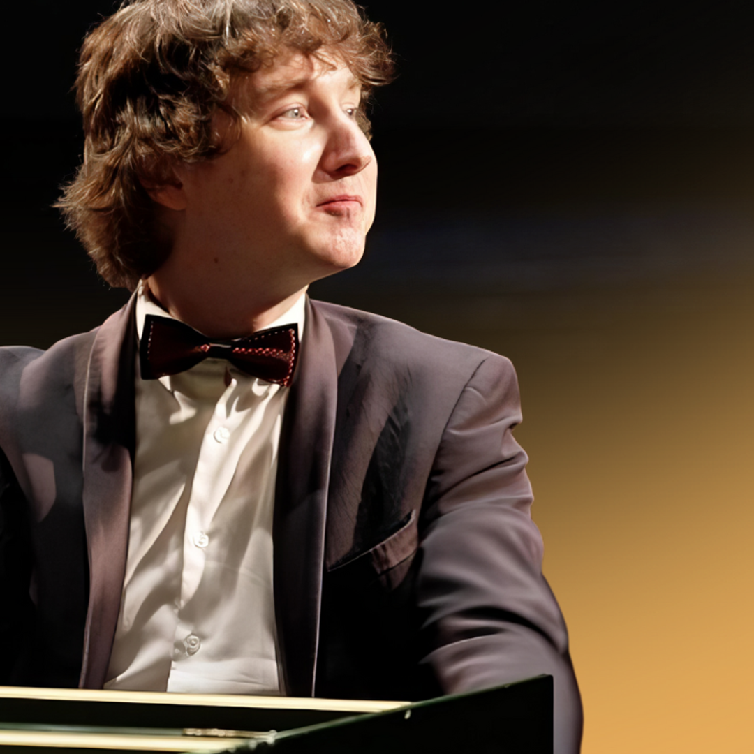 Maxim Emelyanychev sits at the keys. He smiles off to his left. He wears a black suit and bowtie.