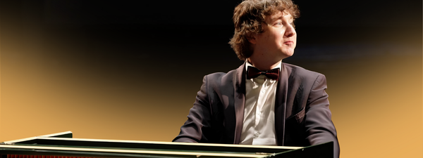 Maxim Emelyanychev sits at the keys. He smiles off to his left. He wears a black suit and bowtie.