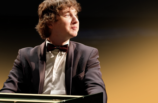 Maxim Emelyanychev sits at the keys. He smiles off to his left. He wears a black suit and bowtie.