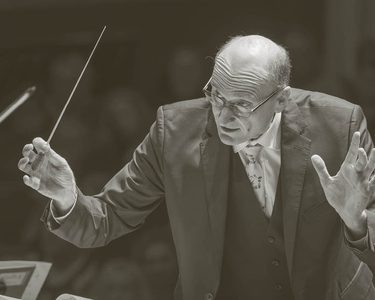 Andrew Manze conducts the Orchestra. He holds a baton and wears a dark suit and glasses.