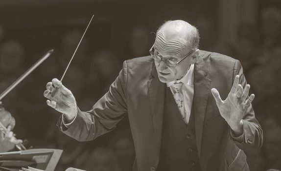 Andrew Manze conducts the Orchestra. He holds a baton and wears a dark suit and glasses.