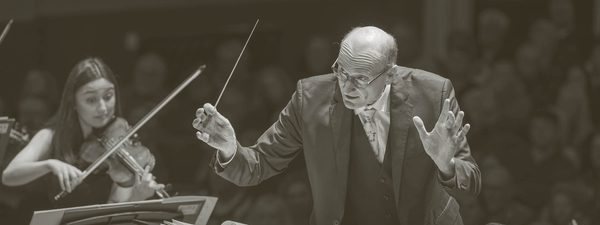 Andrew Manze conducts the Orchestra. He holds a baton and wears a dark suit and glasses.