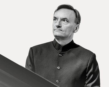 Sir Stephen Hough wears a black shirt and looks off to the right at his piano.