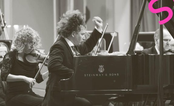 Maxim passionately plays the piano while the orchestra play behind