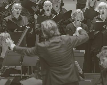 Maxim conducts the orchestra with the chorus singing behind.