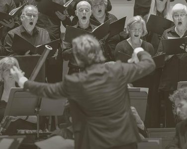 Maxim conducts the orchestra with the chorus singing behind.