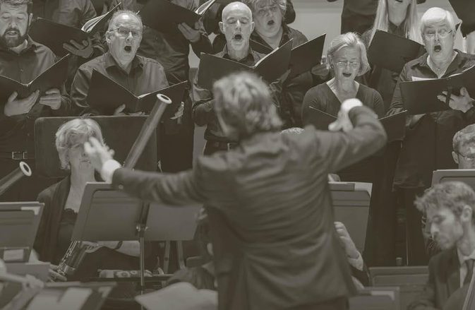Maxim conducts the orchestra with the chorus singing behind.