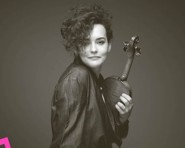Alena smiles and faces the camera with a violin clutched close to her.