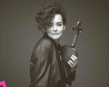 Alena smiles and faces the camera with a violin clutched close to her.