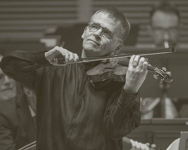 Pekka plays to the audience - the orchestra perform behind him. He wears light glasses and a black long shirt.