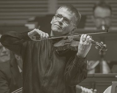 Pekka plays to the audience - the orchestra perform behind him. He wears light glasses and a black long shirt.