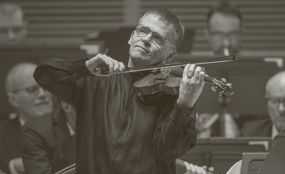 Pekka plays to the audience - the orchestra perform behind him. He wears light glasses and a black long shirt.