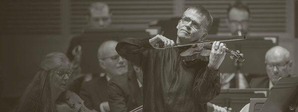 Pekka plays to the audience - the orchestra perform behind him. He wears light glasses and a black long shirt.