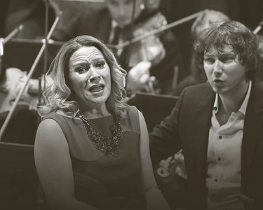 Carolyn Sampson sings and Maxim conducts while the orchestra play behind them. Carolyn wears a sleeveless dress and Maxim wears a dark suit with an unbuttoned white shirt.