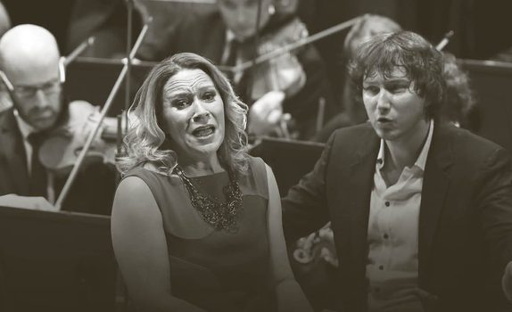 Carolyn Sampson sings and Maxim conducts while the orchestra play behind them. Carolyn wears a sleeveless dress and Maxim wears a dark suit with an unbuttoned white shirt.