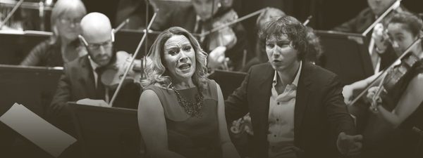 Carolyn Sampson sings and Maxim conducts while the orchestra play behind them. Carolyn wears a sleeveless dress and Maxim wears a dark suit with an unbuttoned white shirt.