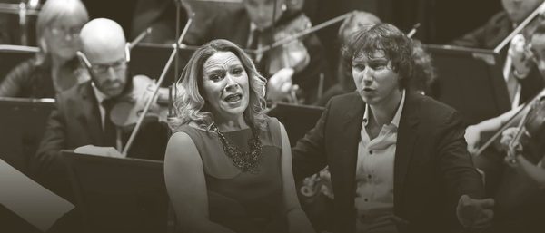 Carolyn Sampson sings and Maxim conducts while the orchestra play behind them. Carolyn wears a sleeveless dress and Maxim wears a dark suit with an unbuttoned white shirt.