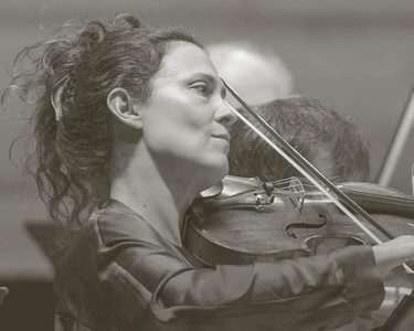Lorenza Borrani plays the violin while the orchestra plays behind.